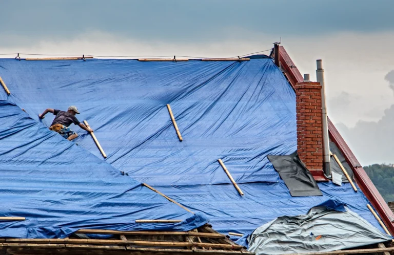 how to tarp a roof-a person using wooden boards to secure a blue tarp over a roof