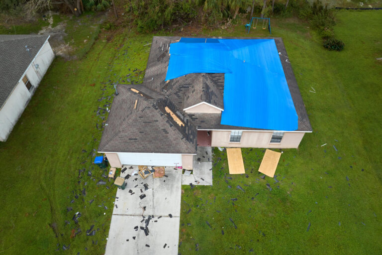 roof warranty-arial view of house surrounded by debris and the roof partially covered in a blue tarp